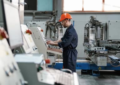 Pracownik ubrany w kask i kombinezon ochronny obsługuje panel sterowania w zakładzie produkcyjnym.