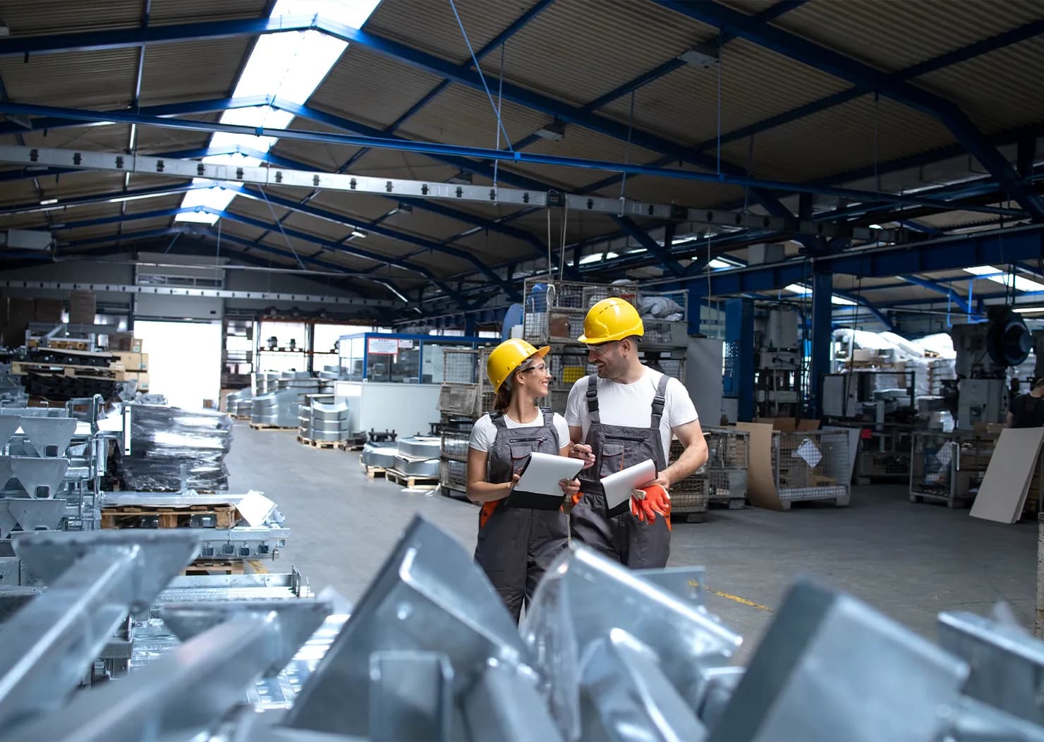 Dwie osoby w kaskach stoją w fabryce, otoczone maszynami i sprzętem przemysłowym.