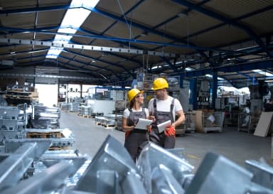 Dwie osoby w kaskach stoją w fabryce, otoczone maszynami i sprzętem przemysłowym.